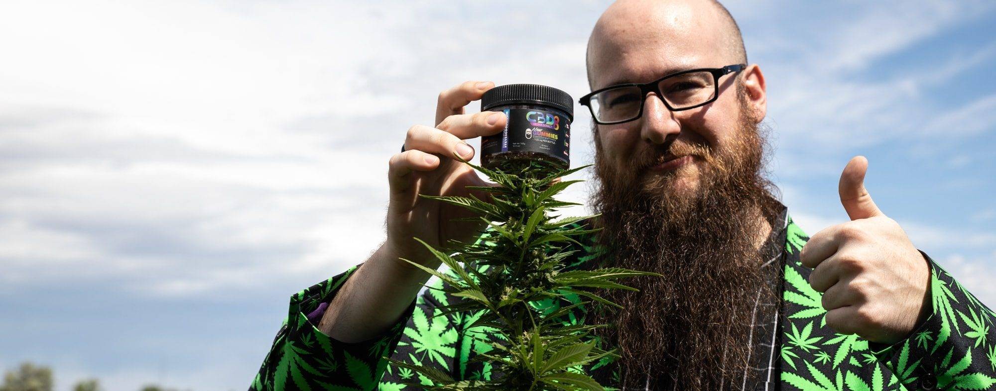 Steve holding CBD edibles in his hand next to a hemp plant in a weed jacket