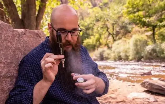 A bearded man smoking a vaporizer near a river.